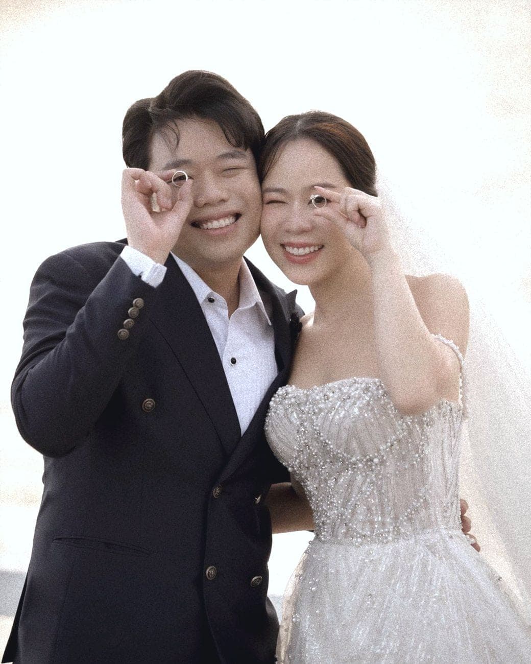 Witita and Sorratat holding their wedding rings up to one eye, by the sea