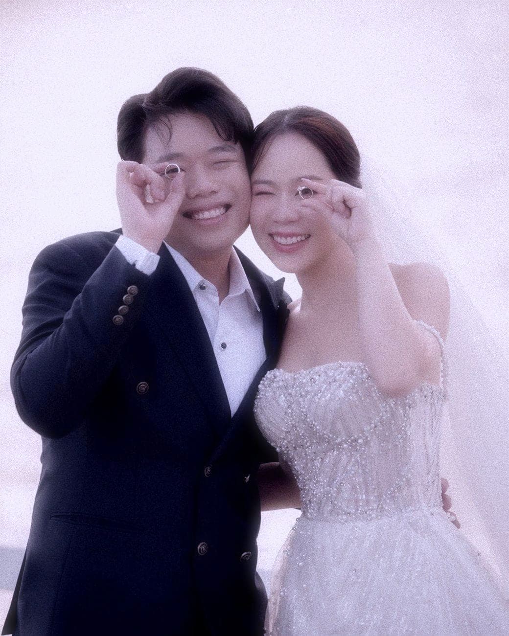 Witita and Sorratat holding their wedding rings up to one eye, by the sea