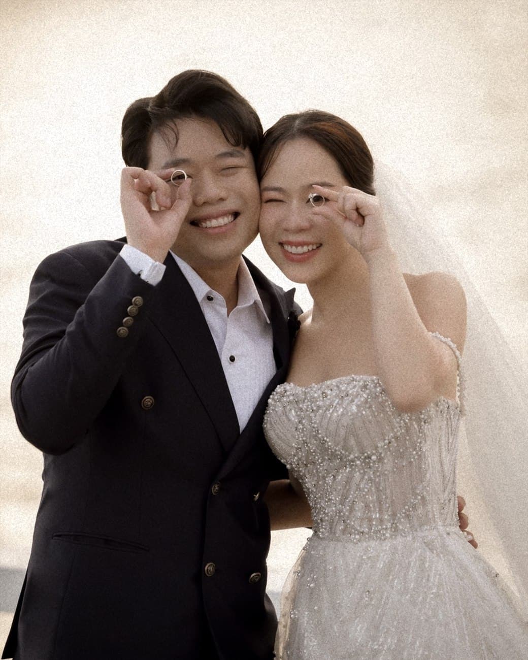 Witita and Sorratat holding their wedding rings up to one eye, by the sea