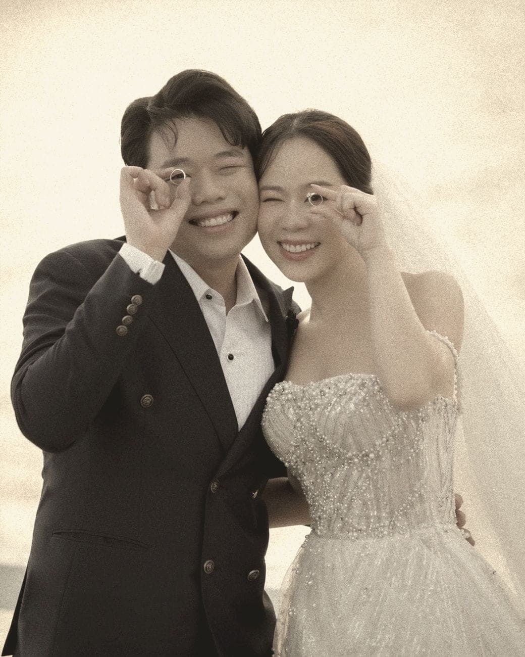 Witita and Sorratat holding their wedding rings up to one eye, by the sea