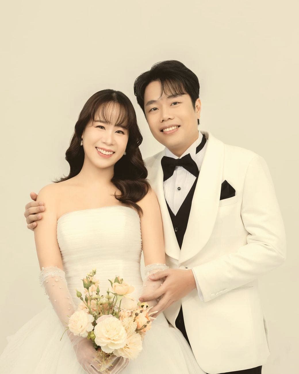Witita holding a bouquet beside Sorratat in a white tuxedo, both smiling