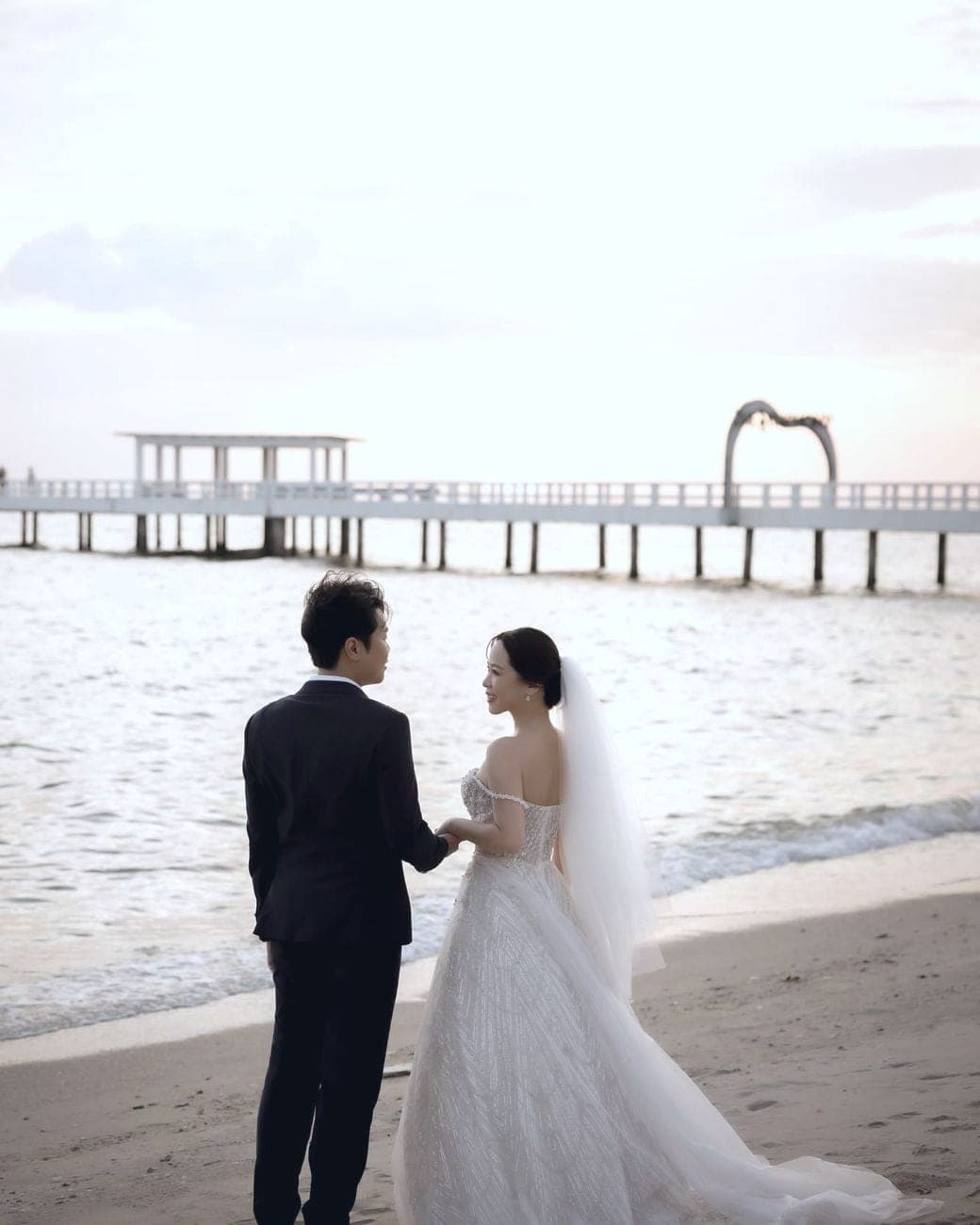 Witita and Sorratat hand in hand on the beach, a long pier behind them